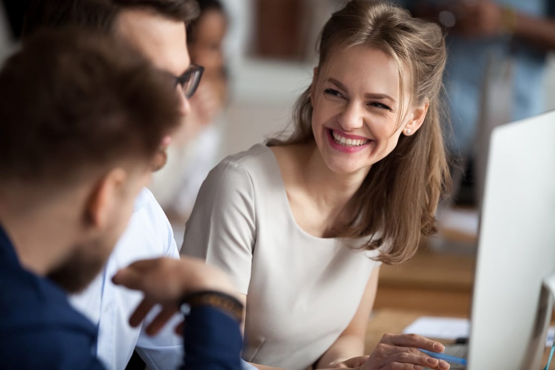 Smiling happy young woman talking with male colleagues at shared workplace, worker laughing at funny joke, reading funny news, excited business success, employees having break, pause